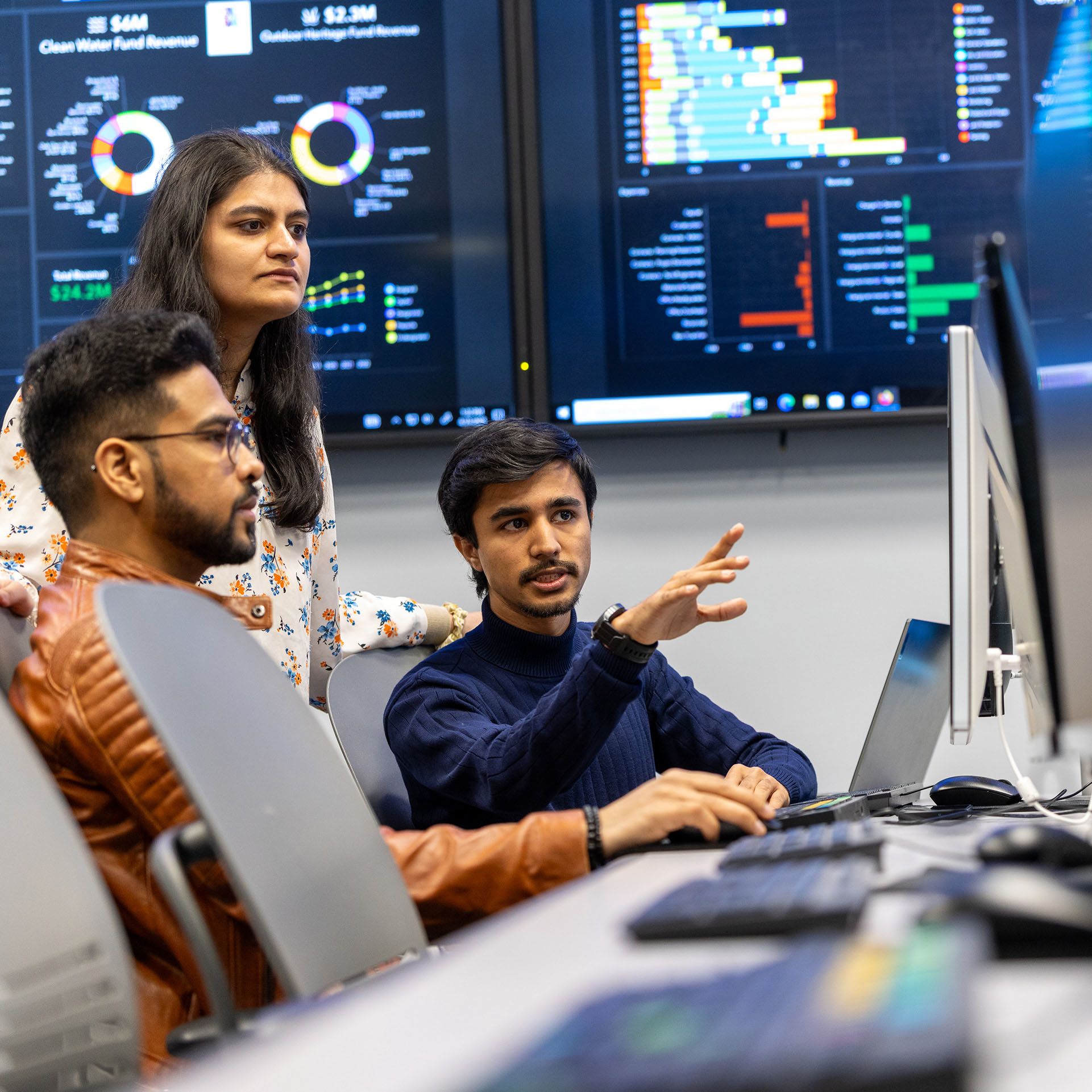 Three students collaborate in a business lab with statistics on monitors behind them.