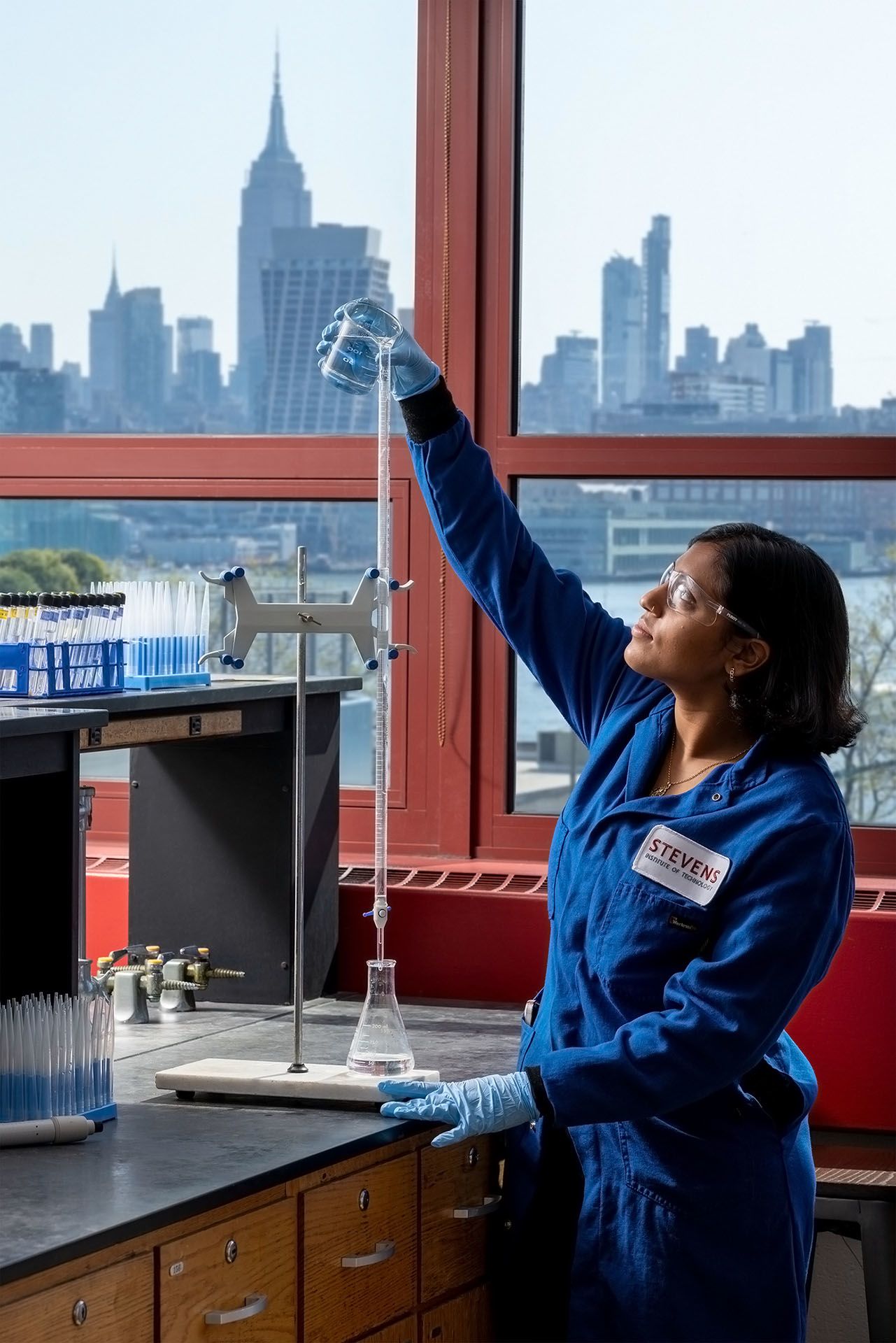 Student in lab coat pours liquid into a beaker in classromm that has a view of the Empire State Building in NYC.