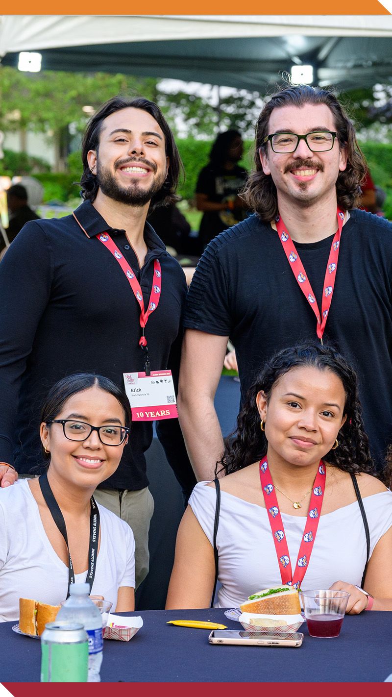 A group of alumni smile for the camera outdoors on campus.